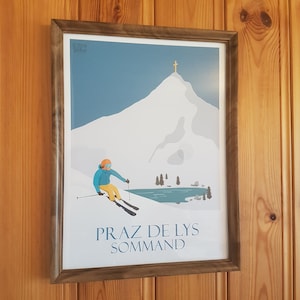 Op de afbeelding: Een ingelijste poster met een blauwe en witte illustratie van een skiër op een besneeuwde berg met een meer op de voorgrond. De berg heeft een kruis op de top. De tekst "PRAZ DE LYS SOMMAND" staat onderaan de poster.