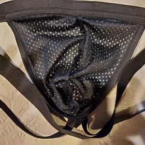 Pode incluir: Jockstrap de malha preta com al&ccedil;as ajust&aacute;veis. As al&ccedil;as s&atilde;o feitas de uma faixa el&aacute;stica larga.