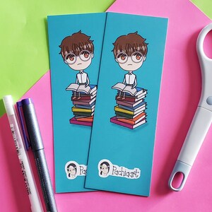 Puede incluir: Dos marcadores de color azul turquesa con una ilustración de dibujos animados de una persona con gafas sentada en una pila de libros. Los marcadores tienen el texto "Pachiqart" en la parte inferior.