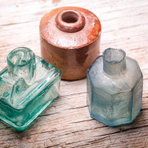 Könnte beinhalten: Drei antike Glasflaschen und ein Keramiktopf mit Deckel auf einer Holzoberfläche. Die Flaschen sind rechteckig und sechseckig, in Blau- und Grüntönen. Der Topf ist braun.