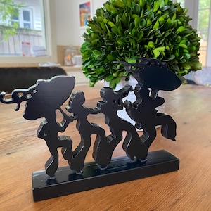 May include: A black wooden sculpture depicting stylized figures in a row, mounted on a rectangular base. The figures have abstract shapes and are connected. The sculpture is placed on a wooden table.