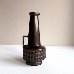 May include: A dark brown ceramic vase with a handle and a textured, patterned base. The vase has a tall, slender neck and a flared top. The handle is rectangular. The vase is approximately 12 inches tall.