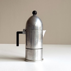 Könnte beinhalten: Ein silberner Espressokocher für den Herd mit einem schwarzen Griff und einem schwarzen Knopf oben. Der Kaffeezubereiter hat eine abgerundete Oberseite und einen Ausguss zum Ausgießen. Der Espressokocher ist aus Metall und steht auf einer weißen Oberfläche.
