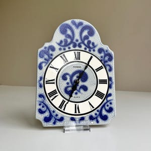 May include: A decorative quartz clock with a white face and black Roman numerals. The clock has a blue and white floral design around the face and on the frame. The clock is supported by a clear stand.