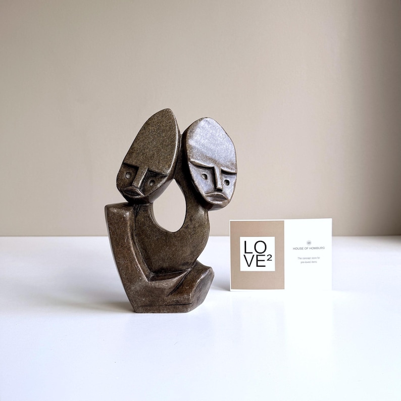 May include: A bronze-colored sculpture depicting two stylized faces joined together. The sculpture has a smooth, polished finish and sits on a white surface. A card with the text "LOVE&sup2;" is next to the sculpture.