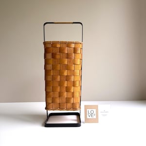 Puede incluir: Una cesta alta y rectangular con cuerpo de chapa de madera marr&oacute;n claro trenzada, sujeta dentro de un marco de metal negro. El asa superior est&aacute; envuelta en un cord&oacute;n color canela. Una peque&ntilde;a tarjeta blanca con el texto LO VE2 est&aacute; al lado.