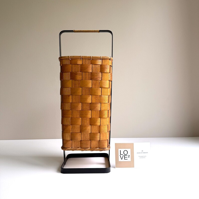 Puede incluir: Una cesta alta y rectangular con cuerpo de chapa de madera marr&oacute;n claro trenzada, sujeta dentro de un marco de metal negro. El asa superior est&aacute; envuelta en un cord&oacute;n color canela. Una peque&ntilde;a tarjeta blanca con el texto LO VE2 est&aacute; al lado.