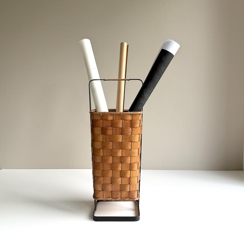 Puede incluir: Una cesta de almacenamiento alta y rectangular con un exterior de madera trenzada de color marr&oacute;n claro y un marco de metal negro. La cesta cuenta con un asa superior y contiene tres tubos de papel enrollados en blanco, beige y negro.