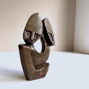 May include: A brown stone sculpture depicting two stylized faces with elongated heads. The sculpture has a smooth, polished finish and a minimalist design. The faces are connected by a curved form, creating a sense of unity. The sculpture is a decorative art piece.