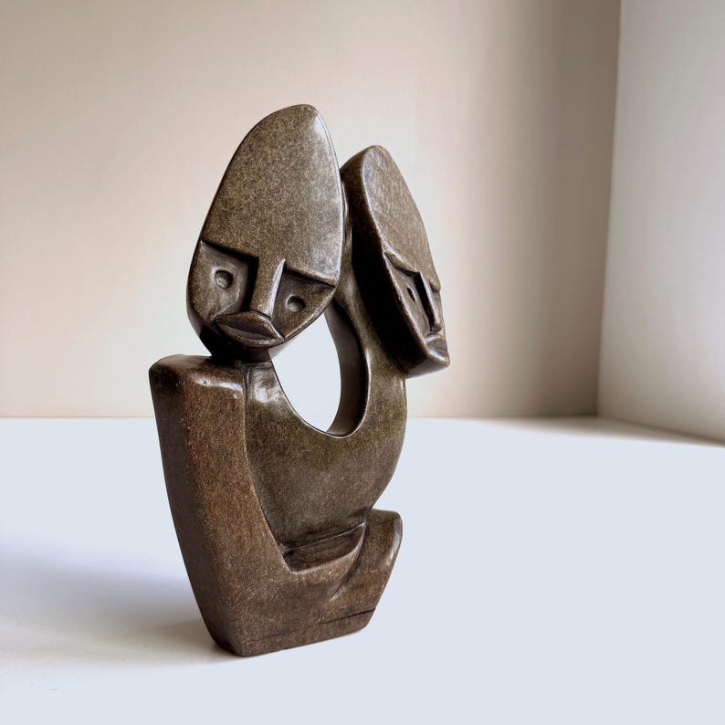 May include: A brown stone sculpture depicting two stylized faces with elongated heads. The sculpture has a smooth, polished finish and a minimalist design. The faces are connected by a curved form, creating a sense of unity. The sculpture is a decorative art piece.