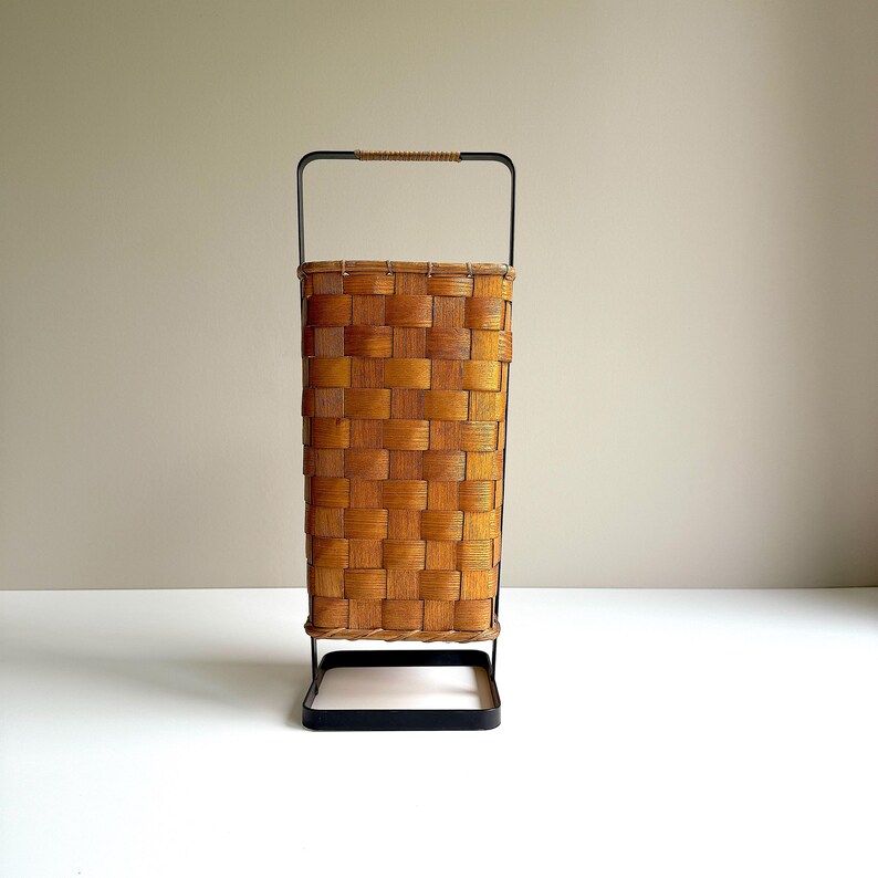 Puede incluir: Una cesta alta y rectangular con un cuerpo de chapa de madera de color marr&oacute;n claro trenzado. La cesta est&aacute; sostenida por un marco de metal negro que se extiende hacia arriba para formar un asa, con una peque&ntilde;a secci&oacute;n de cuerda beige.