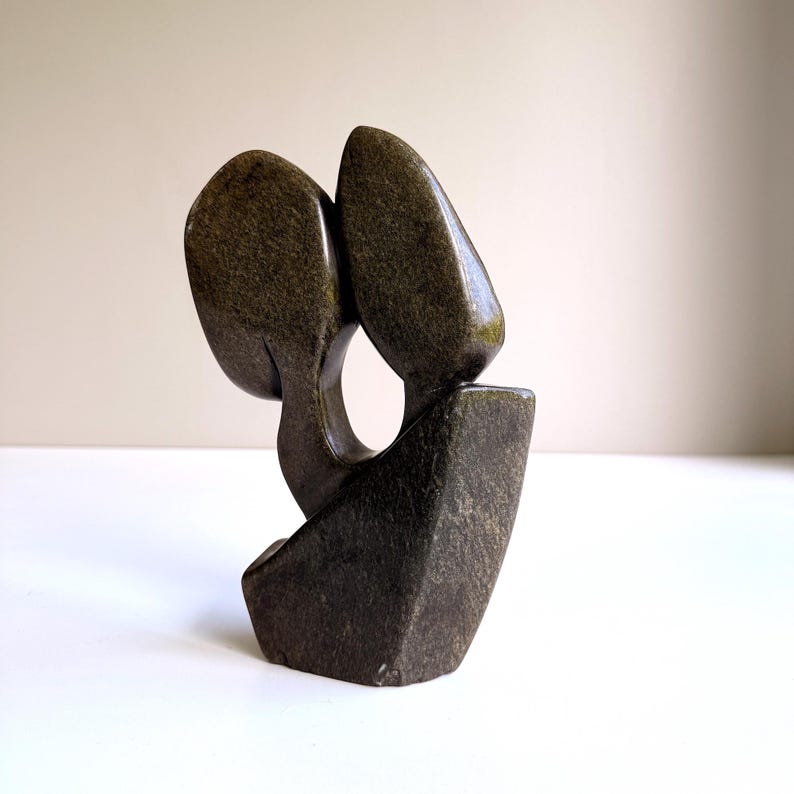 May include: A dark green stone sculpture with a smooth, polished finish. The abstract design features two rounded shapes connected by a curved element, resting on a rectangular base. The sculpture has a natural, earthy aesthetic.