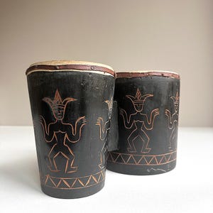 May include: Two black wooden drums with intricate carvings of stylised figures in a reddish-brown colour. The drums are decorated with a geometric pattern around the bottom.