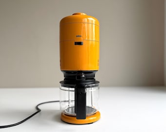 May include: A bright yellow Braun coffee maker with a cylindrical top housing and a clear glass carafe base. The appliance features a black handle on the carafe, a black power cord extending to the left, and a small black power switch on the front.