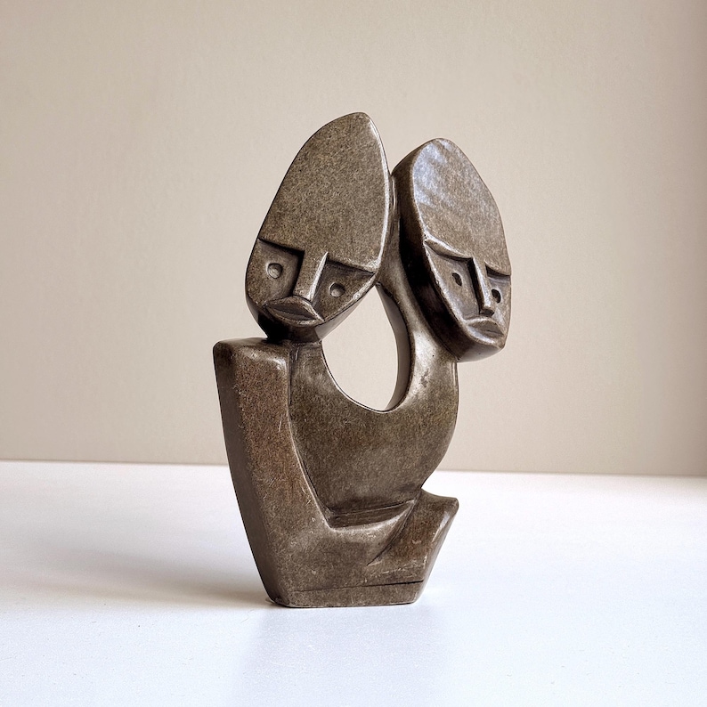 May include: A stone sculpture featuring two stylized faces with elongated heads and simple facial features. The sculpture is a muted brown color and has a smooth, polished finish. The artwork is abstract and modern, with a minimalist design.