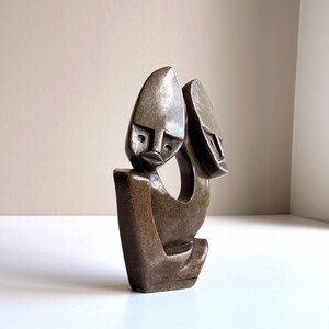 May include: A bronze-toned sculpture depicting two stylized faces. The faces have elongated features and are connected by a curved form. The sculpture has a smooth, polished appearance and sits on a white surface.