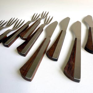 Puede incluir: Un conjunto de cubiertos antiguos, que incluye tenedores y cuchillos. Los utensilios tienen mangos de madera oscura con un acento y hojas de acero inoxidable. Los tenedores tienen cuatro púas. Los cuchillos tienen una hoja redondeada.