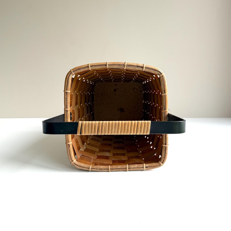 Puede incluir: Una cesta tejida rectangular con acabado en madera marr&oacute;n claro y un asa de metal negro. La cesta presenta un patr&oacute;n de rejilla de tiras de madera finas y una base s&oacute;lida, vista desde arriba para mostrar el espacio interior abierto.