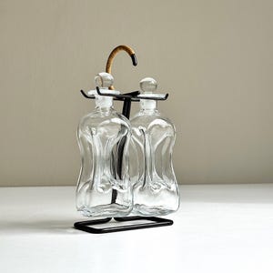 May include: A clear glass cruet set with two bottles, each with a rounded stopper. The bottles are held in a black metal stand with a curved handle wrapped in a light brown material. The bottles have a unique, curved shape.