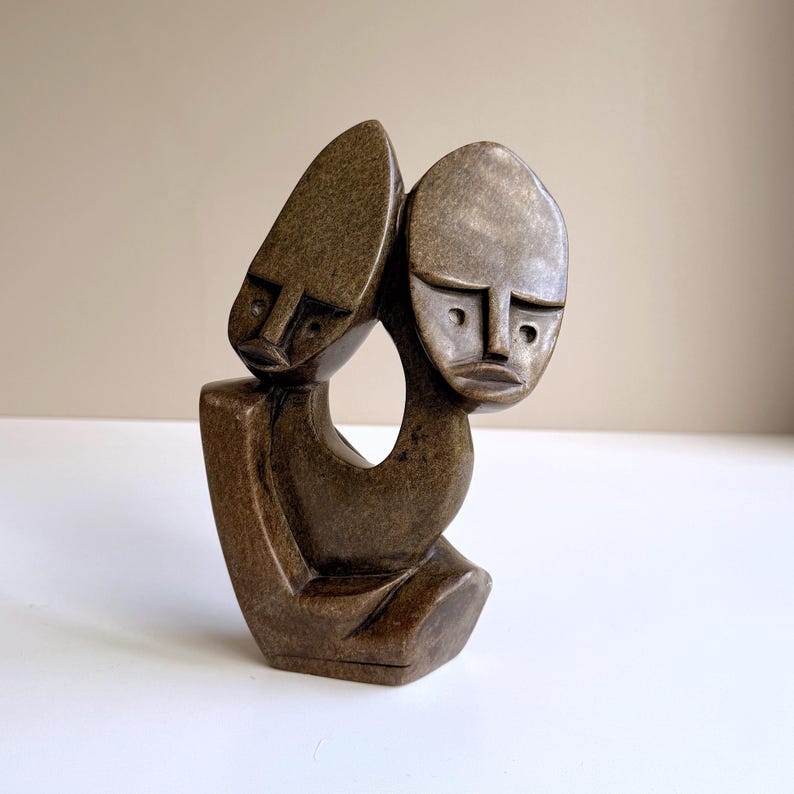 May include: A brown stone sculpture depicting two stylized faces. The faces are oval-shaped with simple features, connected by a curved form. The sculpture has a smooth, polished finish and is set against a neutral background.