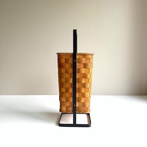 Puede incluir: Una cesta rectangular alta con un patr&oacute;n de tejido de grano de madera marr&oacute;n claro. La cesta est&aacute; sostenida por un marco de metal negro que se extiende hacia arriba para formar un asa vertical en cada lado, asegurada con peque&ntilde;os remaches met&aacute;licos plateados.
