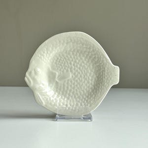 Könnte beinhalten: Eine cremefarbene Keramikschale in Fischform mit einem strukturierten Schuppenmuster auf dem Körper, einem ausgeprägten Kopf mit Auge und einer kleinen Schwanzflosse. Die Schale wird aufrecht auf einem transparenten Acrylständer präsentiert.