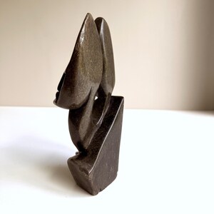 May include: A dark brown stone sculpture of an abstract face. The sculpture has a smooth, polished surface and is mounted on a rectangular base. The face has a prominent nose and a closed mouth. The sculpture is a unique piece of art.