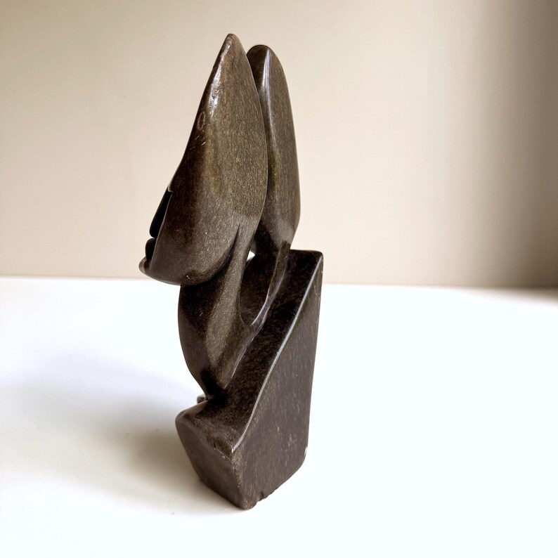 May include: A dark brown stone sculpture of an abstract face. The sculpture has a smooth, polished surface and is mounted on a rectangular base. The face has a prominent nose and a closed mouth. The sculpture is a unique piece of art.
