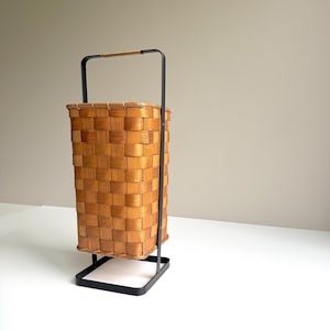 Puede incluir: Una cesta rectangular tejida con un marco y asa de metal negro. La cesta está hecha de material tejido marrón claro. El asa está envuelta con un material marrón claro. La cesta está sobre un soporte de metal negro.