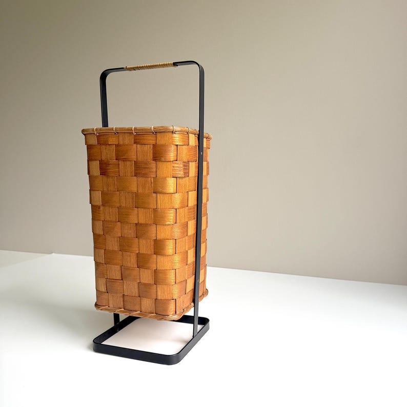 Puede incluir: Una cesta rectangular de madera trenzada con acabado marr&oacute;n claro, suspendida dentro de un marco de metal negro. El marco cuenta con un asa alta con un detalle envuelto en la parte superior y una base cuadrada que sostiene la cesta.