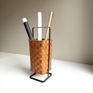 Puede incluir: Una cesta alta y rectangular de madera trenzada con acabado marr&oacute;n claro, sostenida por un marco de metal negro con asa. Tres tubos cil&iacute;ndricos en negro, blanco y beige est&aacute;n colocados verticalmente dentro de la cesta, sobresaliendo.