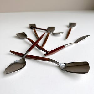 Könnte beinhalten: Ein Set aus sechs silbernen Löffeln mit braunen Holzgriffen. Die Löffel sind kreuzweise auf einer weißen Oberfläche angeordnet.