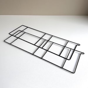 May include: Two black metal wire grid panels, rectangular in shape, are stacked on a white surface. The grids have a geometric design with intersecting lines, creating a modern, minimalist aesthetic. The panels are likely for display or organization.