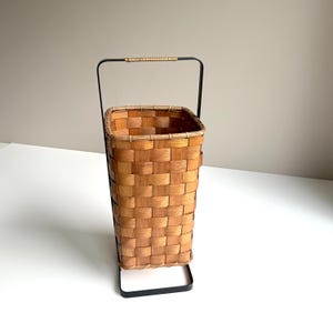 Puede incluir: Una cesta alta y rectangular de madera tejida con un acabado marr&oacute;n claro, que cuenta con un marco de metal negro que se extiende hasta formar un asa con un agarre de rat&aacute;n envuelto en la parte superior. Dise&ntilde;o abierto y estructurado.