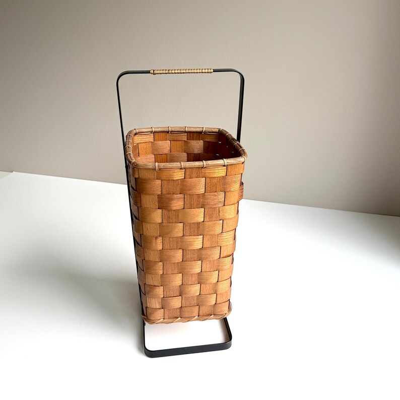 Puede incluir: Una cesta alta y rectangular de madera tejida con un acabado marr&oacute;n claro, que cuenta con un marco de metal negro que se extiende hasta formar un asa con un agarre de rat&aacute;n envuelto en la parte superior. Dise&ntilde;o abierto y estructurado.