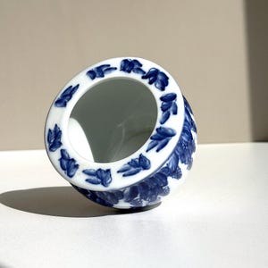Pode incluir: Uma tigela de cerâmica branca e azul com um design floral. A borda é decorada com padrões azuis em forma de folha. O corpo da tigela apresenta um detalhado motivo floral azul. A tigela está sobre um fundo neutro.