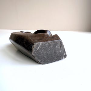 May include: A dark brown, carved stone sculpture with a smooth, rounded top and a rough, textured base. The sculpture has a geometric shape and appears to be a bust or head. The sculpture is set against a neutral background.