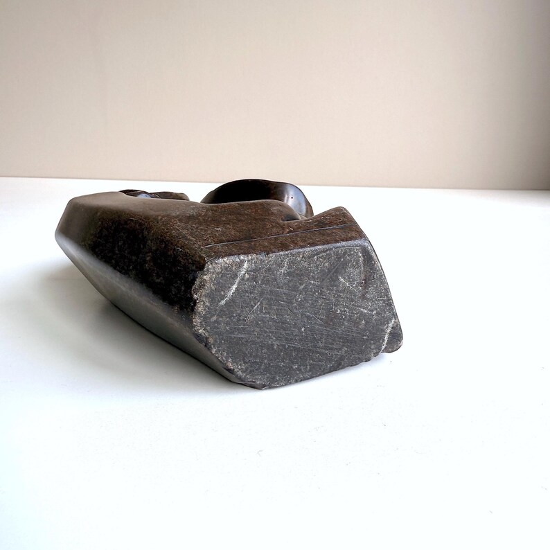 May include: A dark brown, carved stone sculpture with a smooth, rounded top and a rough, textured base. The sculpture has a geometric shape and appears to be a bust or head. The sculpture is set against a neutral background.