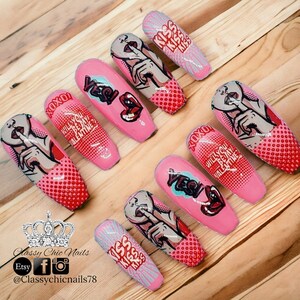 Puede incluir: Una colecci&oacute;n de u&ntilde;as postizas rosas de estilo pop art. Cada u&ntilde;a presenta un dise&ntilde;o diferente, incluyendo labios, la palabra "YES!" y la frase "&iquest;Quieres ser mi San Valent&iacute;n?" Las u&ntilde;as est&aacute;n decoradas con detalles rojos y blancos y un patr&oacute;n de puntos de c&oacute;mic.