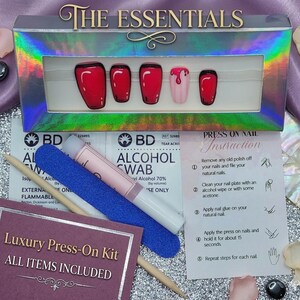 May include: A set of press-on nails in a holographic box labelled THE ESSENTIALS, featuring red nails with black outlines and one nail with a pink drip design. The kit includes an alcohol swab, a blue nail file, a wooden cuticle stick, and instructions.