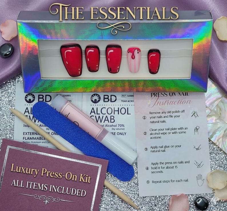 May include: A set of press-on nails in a holographic box labelled THE ESSENTIALS, featuring red nails with black outlines and one nail with a pink drip design. The kit includes an alcohol swab, a blue nail file, a wooden cuticle stick, and instructions.