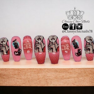 Puede incluir: Un conjunto de diez u&ntilde;as postizas rosas con varios dise&ntilde;os. Algunas u&ntilde;as presentan una mujer de estilo pop art con un dedo en los labios, mientras que otras tienen el texto "KISS" y "WILL YOU BE MY VALENTINE?" Las u&ntilde;as est&aacute;n decoradas con detalles rojos y grises.