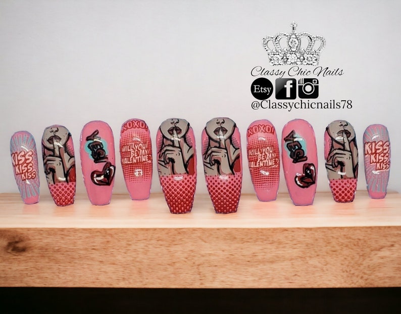 Puede incluir: Un conjunto de diez u&ntilde;as postizas rosas con varios dise&ntilde;os. Algunas u&ntilde;as presentan una mujer de estilo pop art con un dedo en los labios, mientras que otras tienen el texto "KISS" y "WILL YOU BE MY VALENTINE?" Las u&ntilde;as est&aacute;n decoradas con detalles rojos y grises.