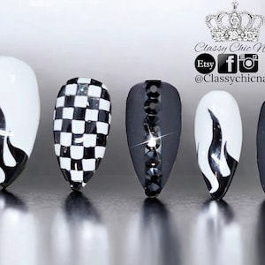 May include: Five press-on nails featuring a black and white color scheme. Designs include white nails with black flame patterns, a black and white checkered pattern, and matte black nails accented with vertical rows of black rhinestones.