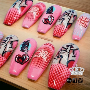 Puede incluir: U&ntilde;as acr&iacute;licas rosas con dise&ntilde;os de arte pop. Las u&ntilde;as presentan im&aacute;genes de labios, corazones y las frases "&iquest;Quieres ser mi San Valent&iacute;n?" y "Kiss Kiss Kiss". Las u&ntilde;as est&aacute;n dispuestas sobre una superficie de madera.