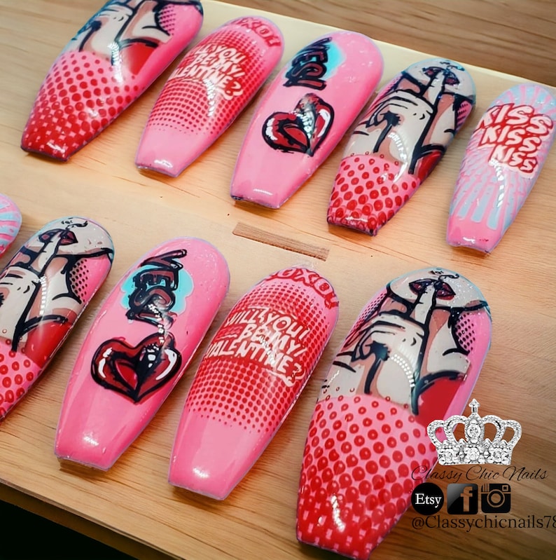 Puede incluir: U&ntilde;as acr&iacute;licas rosas con dise&ntilde;os de arte pop. Las u&ntilde;as presentan im&aacute;genes de labios, corazones y las frases "&iquest;Quieres ser mi San Valent&iacute;n?" y "Kiss Kiss Kiss". Las u&ntilde;as est&aacute;n dispuestas sobre una superficie de madera.