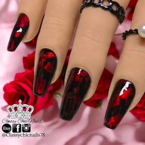 May include: Long coffin-shaped artificial nails featuring a glossy black base with vibrant red foil accents. The nails are displayed on a mannequin hand adorned with a black chain-link ring and a white pearl ring, set against a pink background.