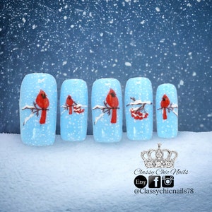 May include: Five light blue nail tips with a winter scene painted on each. Each nail tip features a red cardinal bird perched on a branch with white snow and red berries. The background is a blue sky with white snowflakes.