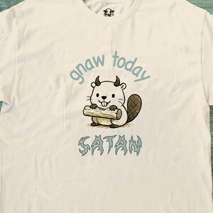 Peut inclure: T-shirt crème avec un dessin animé de castor cornu tenant une bûche. Les mots "gnaw today" sont au-dessus du castor, et "SATAN" est imprimé en dessous dans une police stylisée. Un petit logo est visible près de l'encolure.
