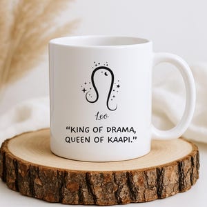 May include: White ceramic mug featuring the Leo zodiac symbol with the text "Leo" and the quote "King of Drama, Queen of Kaapi." The mug has a curved handle and is set on a wooden coaster.